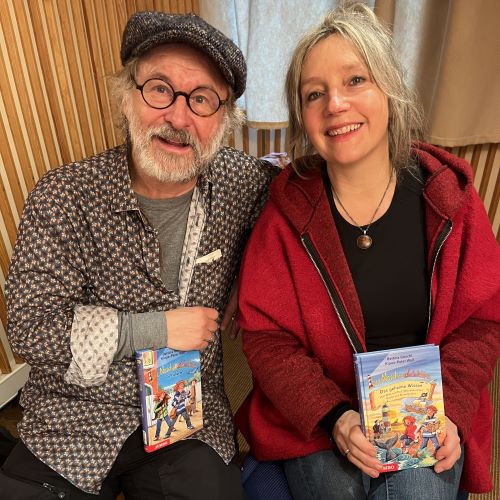 Klaus-Peter Wolf und Bettina Göschl sitzen nebeneinander im Tonstudio. In den Händen halten sie zwei Buchausgaben von 'Die Nordseedetektive'.