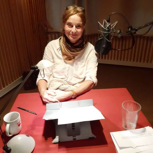 Katrin Gerken sitzt bei der Aufnahme im Tonstudio an einem Tisch. Vor ihr liegt der Text zum Lesen auf einem Ständer. Auf dem Tisch stehen eine Tasse, ein Wasserglas und eine Lampe.